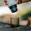 Ръка държи преносима кафемашина Outin Nano над Чаша за еспресо Outin Nano върху дървена повърхност на открито.