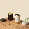 Две чаши Outin Travel Tumblers - черна с рисунка на лате и бяла с кафе, без капаци - са разположени сред зърна върху бежова повърхност. Две чаши Outin Travel Tumblers - черна с рисунка на лате и бяла с кафе, без капаци - са разположени сред зърна върху бежова повърхност.